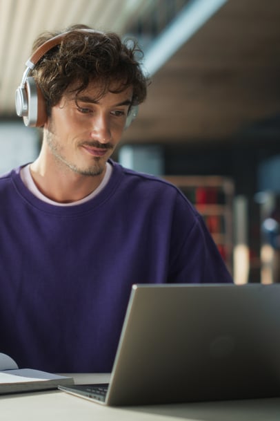 A young man wearing headphones and a purple sweatshirt sits at a table using a laptop.