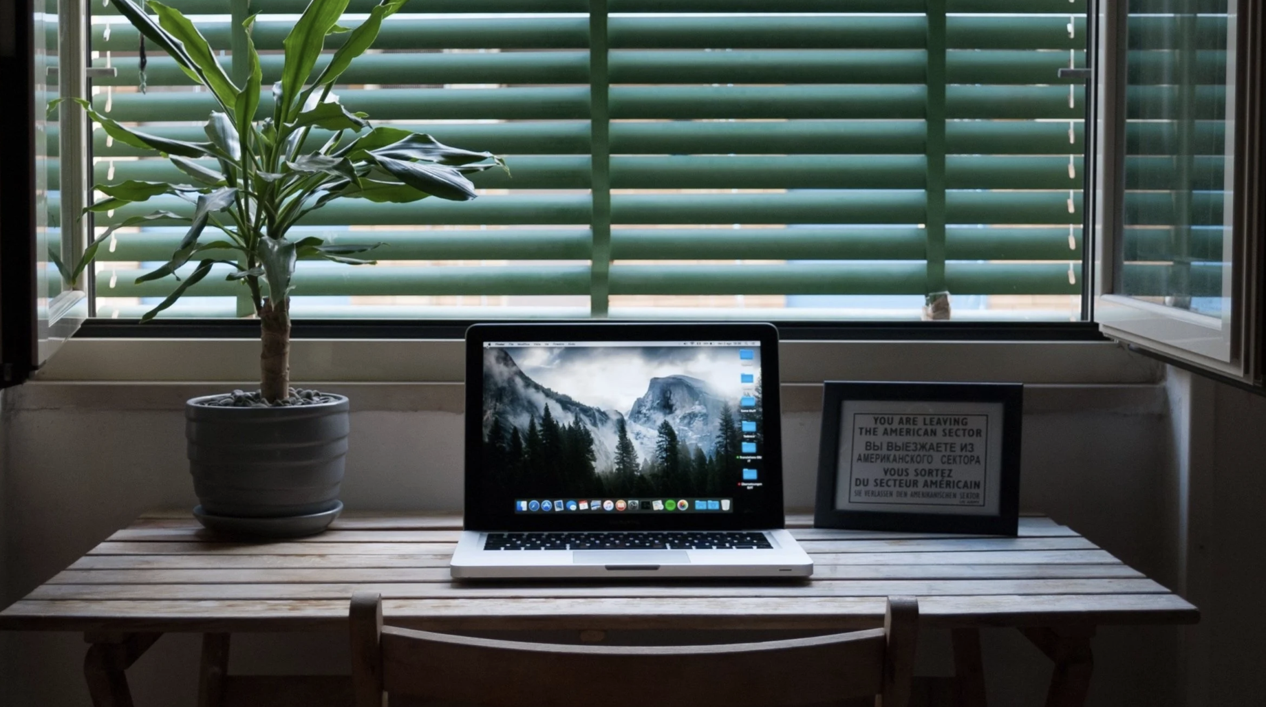 A laptop on a wooden table with a plant and shuttered windows