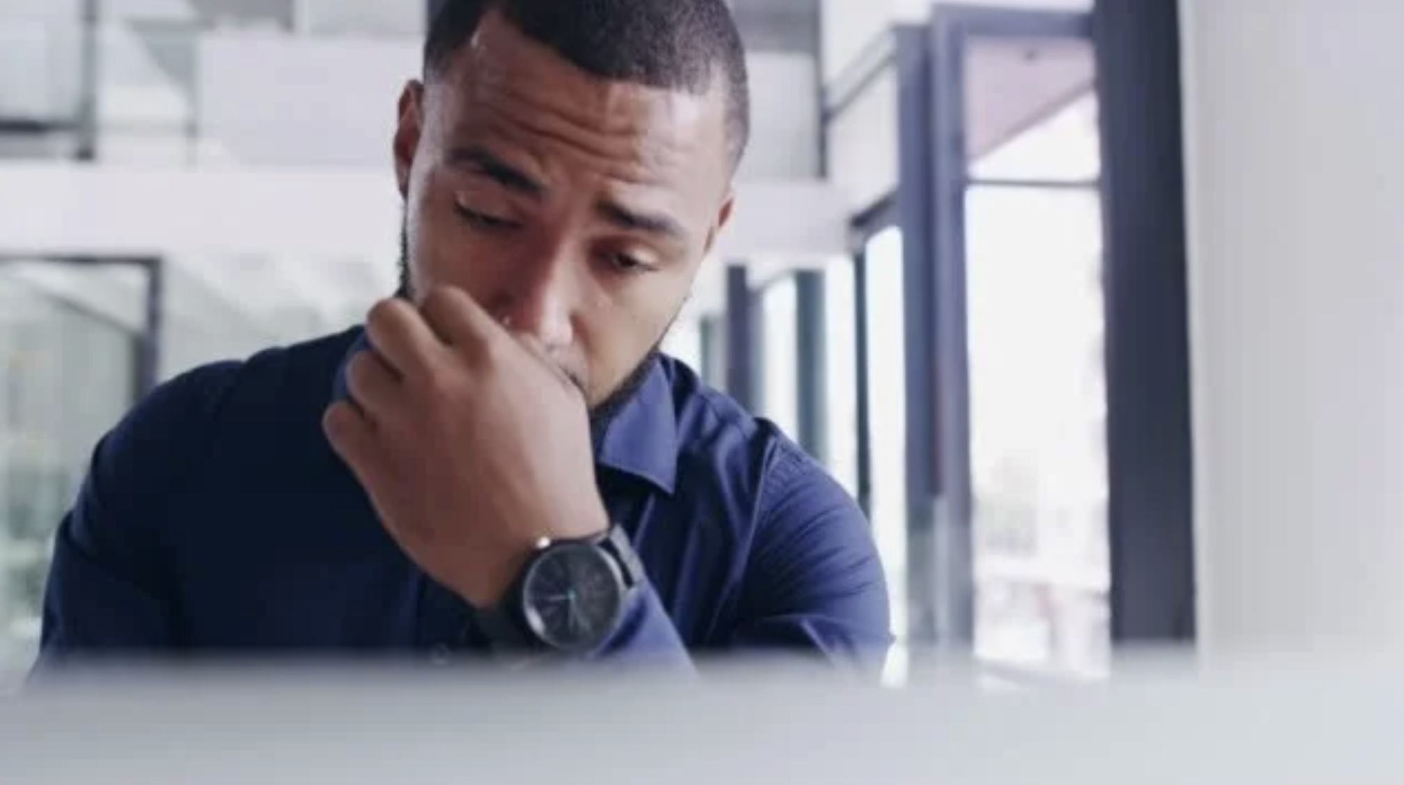 The image shows a person sitting at a desk, appearing to be in a moment of deep thought or concern. The individual is wearing a dark-colored shirt and has a watch on their left wrist. The background suggests an indoor setting with large windows that allow natural light to enter the space.