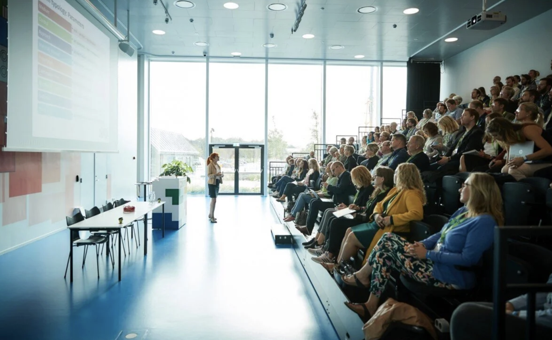 The image captures a moment in a modern conference room bathed in natural light. On the left, a presenter stands at a white podium, gesturing towards a large screen that displays a list of points.