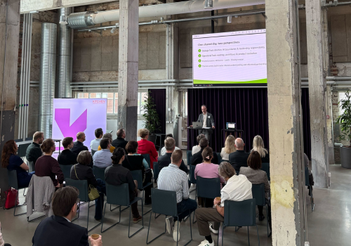 The image shows people sitting on chairs, looking at a man presenting in front of a screen
