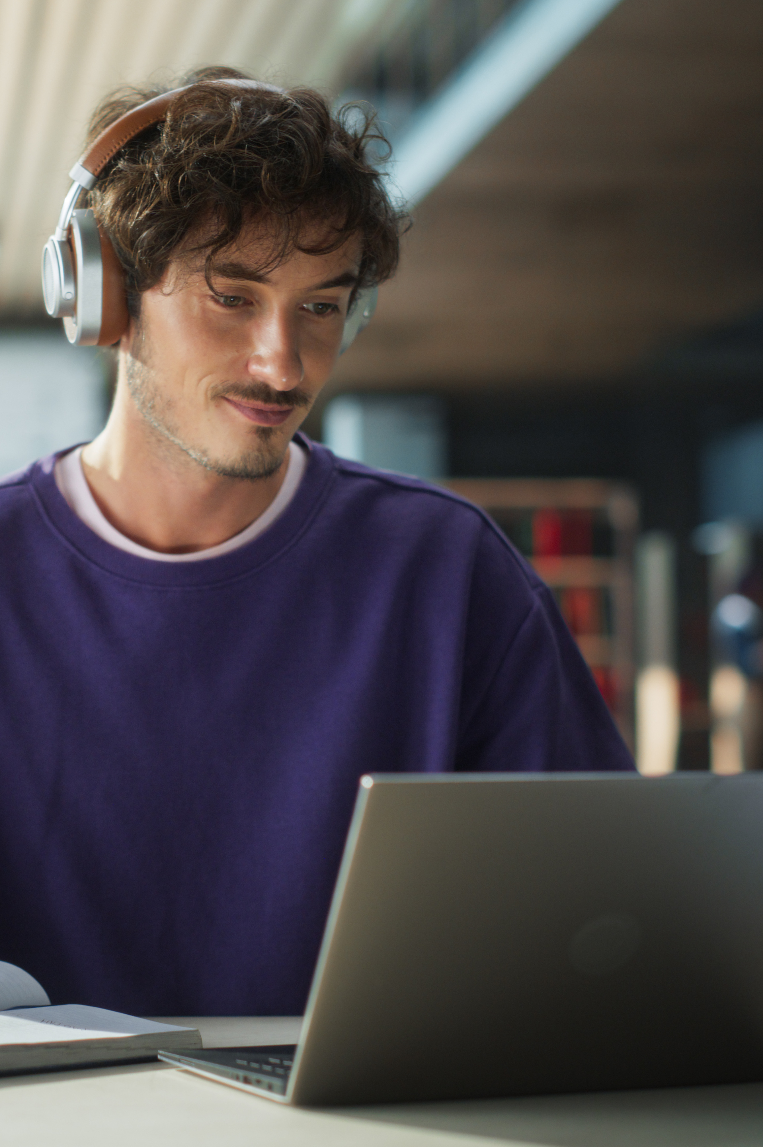 A young man wearing headphones and a purple sweatshirt sits at a table using a laptop.
