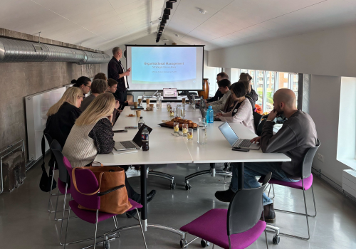 The image shows a group of people sitting around a table, listening to a man presenting in front of a screen
