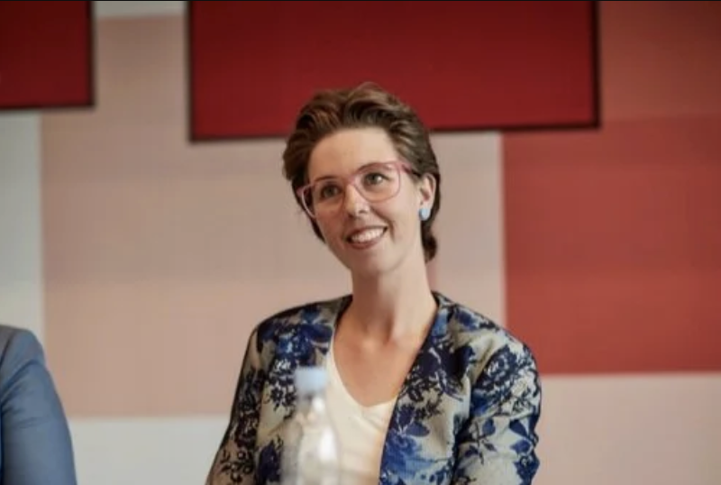 The image shows a woman with short hair, wearing a floral blazer over a light-colored top. She has glasses on and is smiling. The background includes a red wall with two red panels.