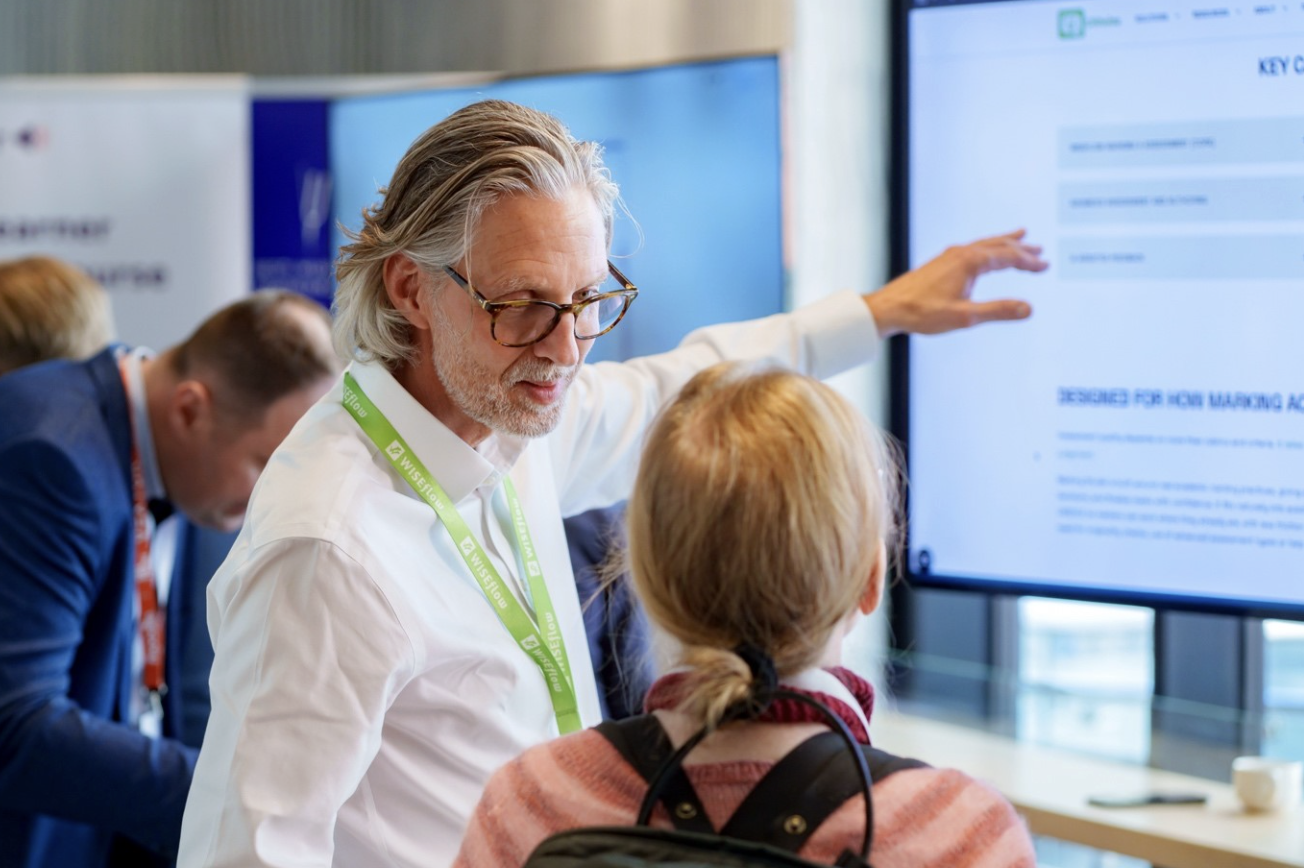 The image captures a moment at an event where Rasmus Blok is pointing at a computer screen while discussing something with a person whose back is to the camera. They appear to be engaged in a presentation or training session, as indicated by the screen content and their focused expressions.